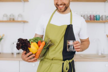 Mutfakta elinde bir tabak taze sebze ve bir bardak su tutan bir adam.