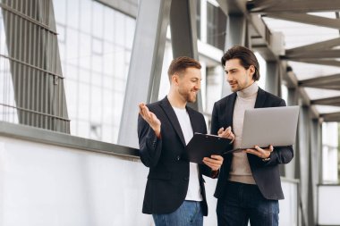Takım elbiseli iki yakışıklı genç iş adamı bir dizüstü bilgisayar ve bir klasör tutuyorlar ve bir çalışma projesini tartışıyorlar ya da kentsel ofis binalarının arka planına karşı video görüşmesi yapıyorlar..