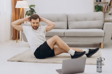 Pozitif atletik sakallı orta yaşlı bir adam evde esniyor, dizüstü bilgisayar kullanıyor, internette spor videoları izliyor, fitness dersi alıyor, kopya çekiyor. Sağlıklı yaşam tarzı, kendini soyutlama sporu.