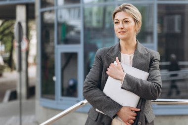 Olgun bir iş kadınının portresi. Duygularıyla gülümseyen ve modern şehir ve ofis binalarında dizüstü bilgisayar tutan.
