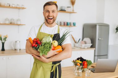 Evdeki mutfakta farklı taze sebzeler tutan mutlu bir adam.