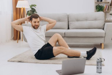 Pozitif atletik sakallı orta yaşlı bir adam evde esniyor, dizüstü bilgisayar kullanıyor, internette spor videoları izliyor, fitness dersi alıyor, kopya çekiyor. Sağlıklı yaşam tarzı, kendini soyutlama sporu.