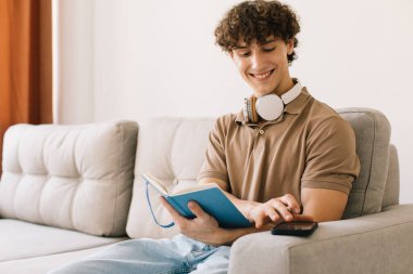 Genç ve çekici kıvırcık saçlı erkek öğrenci oturma odasında kanepede otururken ders çalışıyor.