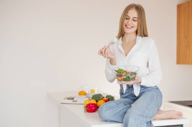 Mutlu ve gülümseyen sarışın kadın mutfaktaki masanın üstünde oturmuş yemek malzemelerinin arasında ev yapımı salatanın tadını çıkarıyor..