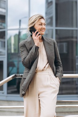 Takım elbiseli, gri ceketli, modern şehir ve ofis binalarında gülümseyen ve konuşan güzel bir iş kadınının portresi.