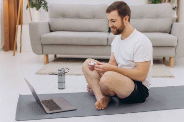 Çekici sakallı adam dizüstü bilgisayarın yanında kablosuz kulaklık tutuyor ve sabah egzersizi ya da yoga yapıyor..