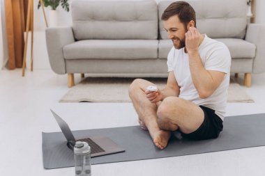 Çekici sakallı adam dizüstü bilgisayarın yanında kablosuz kulaklık tutuyor ve sabah egzersizi ya da yoga yapıyor..
