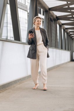 Takım elbiseli, gri ceketli, modern şehir ve ofis binalarında gülümseyen ve konuşan güzel bir iş kadınının portresi.