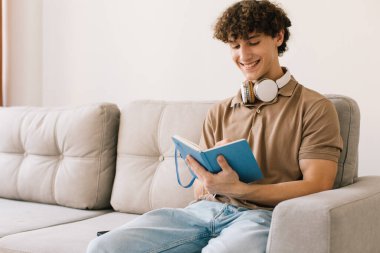 Genç ve çekici kıvırcık saçlı erkek öğrenci oturma odasında kanepede otururken ders çalışıyor.