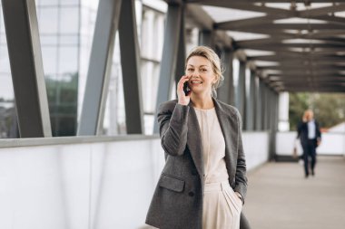 Takım elbiseli, gri ceketli, modern şehir ve ofis binalarında gülümseyen ve konuşan güzel bir iş kadınının portresi.