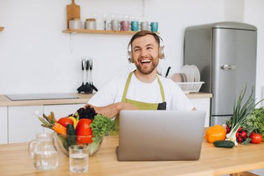 Kulaklıklı mutlu adam mutfakta salata hazırlıyor taze sebzelerden ve dizüstü bilgisayara bakıyor..