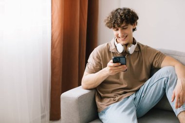 Oturma odasında kanepede oturup dinlenirken akıllı telefon kullanan mutlu kıvırcık saçlı genç adamın portresi.