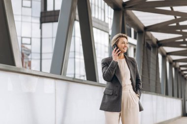 Takım elbiseli, gri ceketli, modern şehir ve ofis binalarında gülümseyen ve konuşan güzel bir iş kadınının portresi.