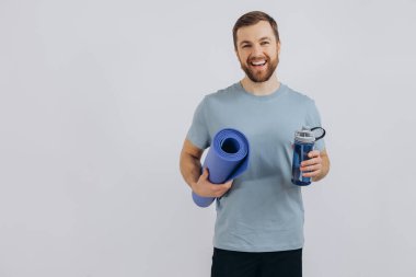 Spor kıyafetli yakışıklı sakallı adamın portresi. Elinde yoga minderi ve beyaz arka planda bir şişe temiz su var.