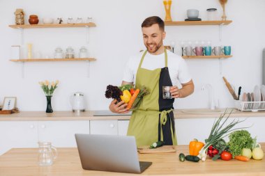 Evde mutlu bir adamın portresi, sebze salatası yapan bir adam kameraya bakıyor ve gülümsüyor, sebzeleri dilimliyor, dizüstü bilgisayarla çevrimiçi pişirme eğitimi alıyor.