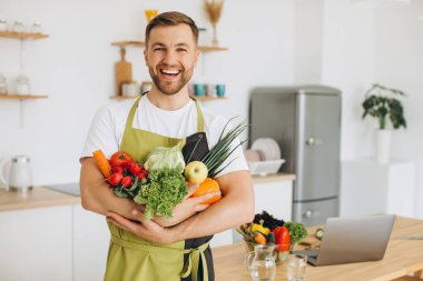 Evdeki mutfakta farklı taze sebzeler tutan mutlu bir adam.