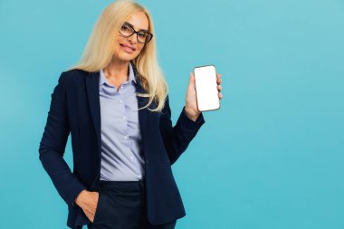 Resmi takım elbiseli, gülümseyen olgun bir iş kadınının portresi. Elinde beyaz, boş bir cihaz ekranı olan akıllı bir telefon, kameraya yakın, mavi stüdyo. Model için boş alanı olan Gadget