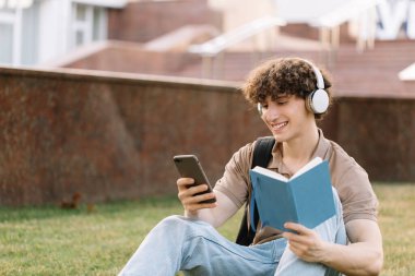 Kulaklıklı çekici erkek öğrenci güneşli bir günde çimlerin üzerinde oturmuş üniversitenin yakınında ödev yapıyor.
