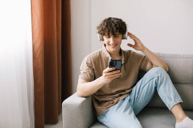 Oturma odasında kanepede oturup dinlenirken akıllı telefon kullanan kıvırcık saçlı genç adamın portresi.