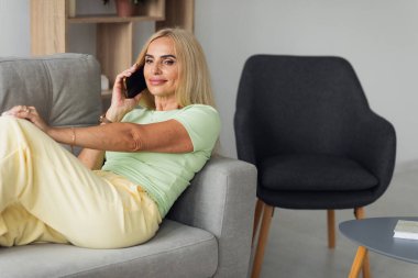 Çekici sarışın orta yaşlı kadın evdeki oturma odasında kanepede otururken telefon kullanıyor.