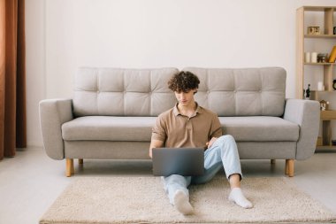 Evdeki kanepede otururken dizüstü bilgisayar kullanan çekici kıvırcık saçlı adamın portresi.