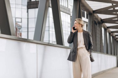 Takım elbiseli, gri ceketli, modern şehir ve ofis binalarında gülümseyen ve konuşan güzel bir iş kadınının portresi.