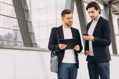 İki modern mutlu iş adamı ellerinde telefon ve dosya ile şehir ofislerinin ve binaların arka planına karşı bir şeyler tartışıyorlar.