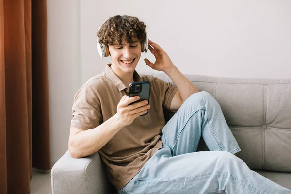 소파에 앉아서 헤드폰을 스마트폰을 사용하는 곱슬머리의 남자의 — 스톡 사진