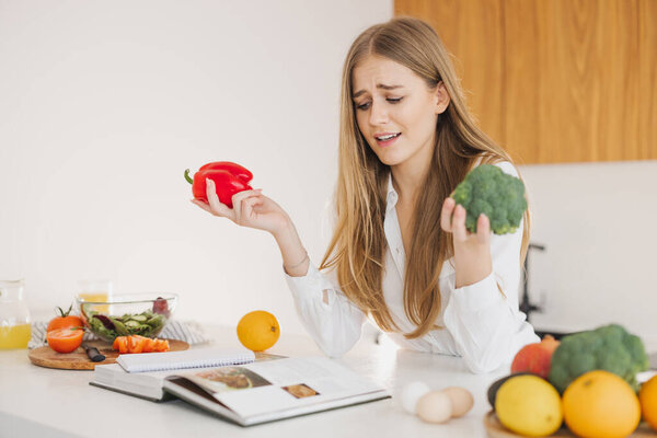Portrait of a smiling cute blonde girl holding bell pepper and broccoli and reading recipe book in kitchen on table among cooking ingredients.
