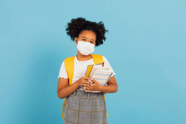 stock image African american schoolgirl in mask holding books on blue background with copy space. Back to school concept. COVID-19