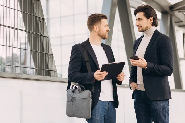 stock image Two modern happy businessmen with phone and folder of documents discussing something against the background of urban offices and buildings