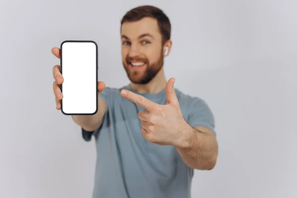 あなたを指しているカメラに白い画面を示す手に空白の携帯電話を保持している陽気な感情的なひげの男 幸せな千年紀の男新しいアプリケーションやモバイルウェブサイトをお勧めします モックアップバナー — ストック写真