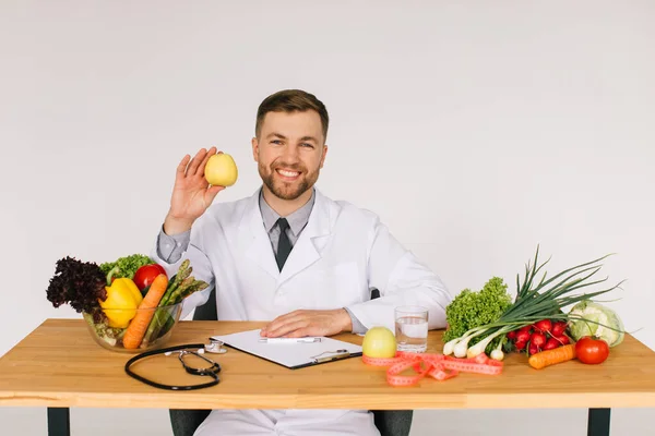 Happy doctor nutritionist sitting at workplace table among fresh vegetables and holding apple