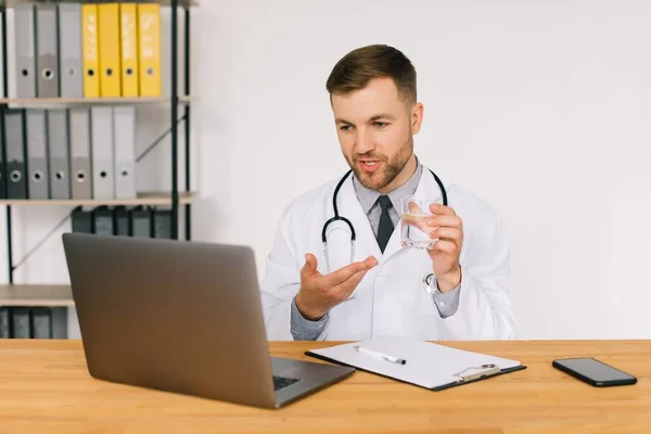 ラップトップで患者とビデオ通話を持つ水のガラスを示す男性医師 オフィスの職場に座っています — ストック写真