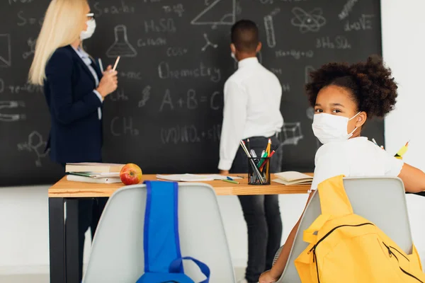 Portrait of African American school girl in mask sitting at desk on a blackboard background in a classroom during a lesson. Mask mode, quarantine, covid-19.