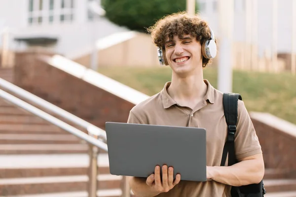 Concept of e-learning, distance study or remote learning concept, Young happy curly hair school guy, college or university student with backpack and headphones using laptop standing on steps in university