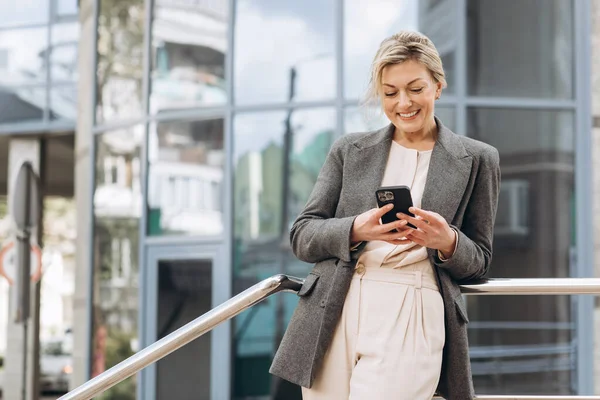 一个穿着西服和灰色夹克的漂亮而成熟的女商人的画像 在现代城市和办公大楼的背景下微笑着 用电话交谈着 — 图库照片
