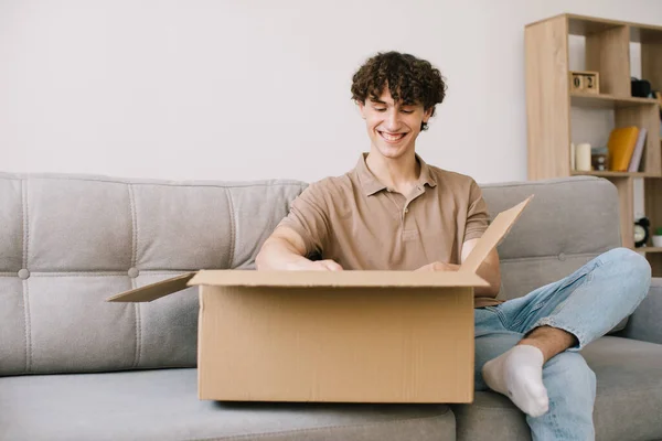 Happy young smiling curly man opening box with ordered goods gifts, presents at home on couch. Online shopper male customer opening online shop parcel.