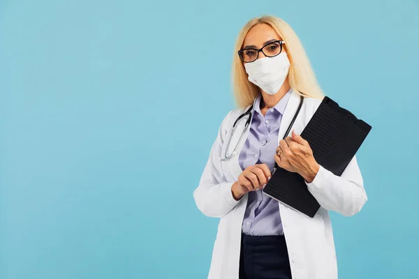 Mature woman doctor in mask holds folder on blue background. Covid-19 concept