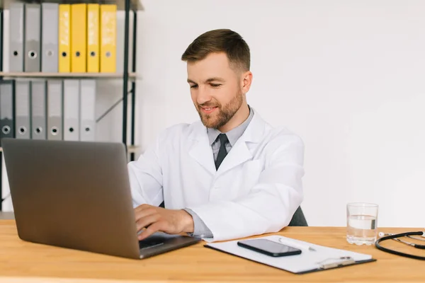 Happy male doctor watching an online medical webinar or training seminar while sitting with a laptop in the workplace.