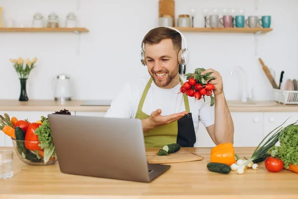 stock image Happy man in headphones sitting at kitchen table and preparing salad, holding radish and showing it to laptop.