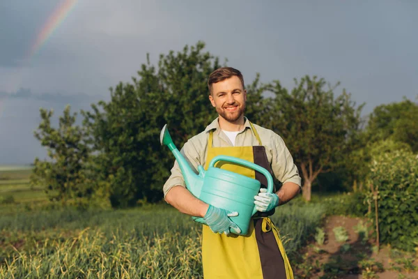 快乐的男性农民在花园里拿着一个浇水罐 背靠着雨后的夏色彩虹 — 图库照片