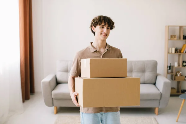 Shopping And Delivery. Curly Young Man Customer Holding Cardboard Box Posing Smiling To Camera Standing At Home. Excited Male Buyer Hugging Delivered Parcel.