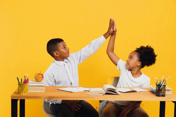 stock image Happy african american schoolgirl and mixed race schoolboy sitting together at desk and giving high five to each other on yellow background. Back to school concept.