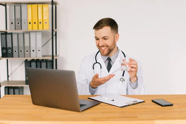 ラップトップで患者とビデオ通話を持つ水のガラスを示す男性医師 オフィスの職場に座っています — ストック写真