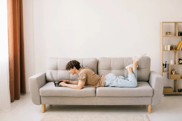 Retrato Atraente Jovem Cabelo Encaracolado Homem Deitado Sofá Usando Laptop — Fotografia de Stock