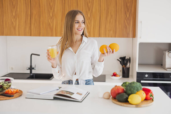 A happy blonde woman is looking at a recipe book and drinking juice in the kitchen