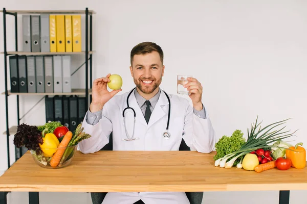 Happy doctor nutritionist sitting at workplace at desk in office among fresh vegetables holding apple and water, diet plan concept