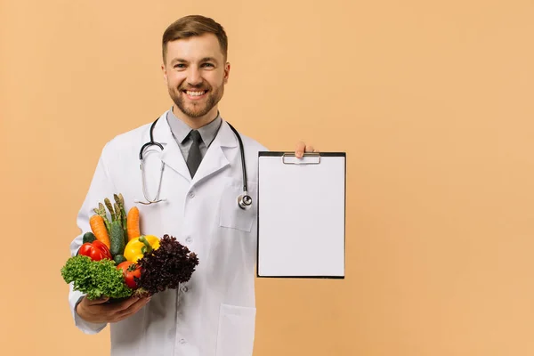 新鮮な野菜やフォルダを保持している聴診器を持つ男性医師栄養士とともにコピースペースでベージュの背景 — ストック写真