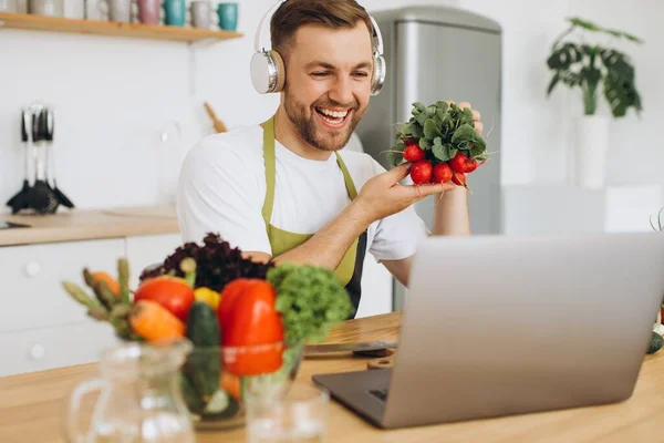 stock image Happy man in headphones sitting at kitchen table and preparing salad, holding radish and showing it to laptop.